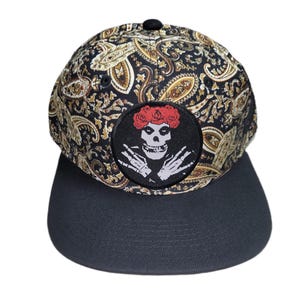 May include: A baseball cap featuring a black and gold paisley pattern on the crown and a solid black flat brim. A circular patch on the front displays a white skull graphic with red flowers on top and skeletal hands positioned below the chin.