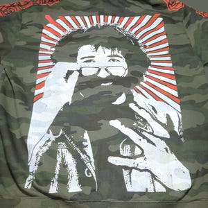 May include: Camouflage-patterned hoodie featuring a white graphic of a man with glasses, a beard, and a peace sign hand gesture. The graphic is set against a red and white burst design. The sleeves have a red floral design.