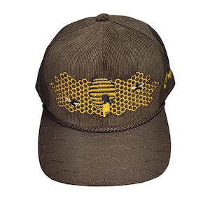 Op de afbeelding: Een bruine corduroy baseballcap met een honingraat- en bijenontwerp geborduurd op de voorkant. De cap heeft een gebogen klep en een gestikt patroon op de kroon. Het ontwerp toont een gele honingraat, een bijenkorf en twee bijen.