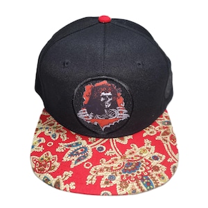 May include: A black baseball cap with a flat brim featuring a red and multi-colored paisley pattern. The front panel displays a circular patch showing a skeletal figure emerging from a torn opening, and the cap is topped with a small red button.