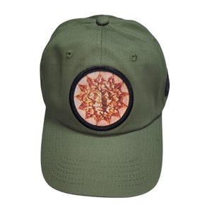 May include: Olive green baseball cap with a circular patch featuring a sun-like design in shades of orange and red. The cap has a curved brim and a button on top. A stylish accessory for everyday wear.