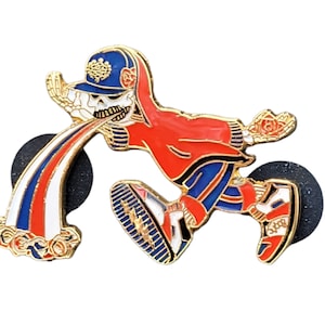 May include: A gold-colored enamel pin of a skeleton wearing a red and blue outfit, a blue baseball cap, and white sneakers. The skeleton is spitting out a rainbow-colored stream of liquid.