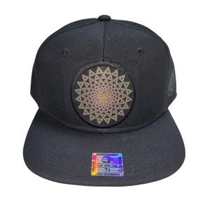 Peut inclure: Casquette noire à visière plate ornée d'un écusson circulaire avec un motif géométrique doré, rose et beige. La casquette a une couronne structurée et une visière plate. Un autocollant holographique est fixé sur la visière.