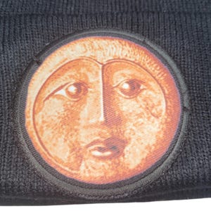 May include: Black knit beanie featuring a circular patch with a stylized face design in shades of orange and brown. The patch is outlined with a black border. The beanie is made of a soft, textured material.