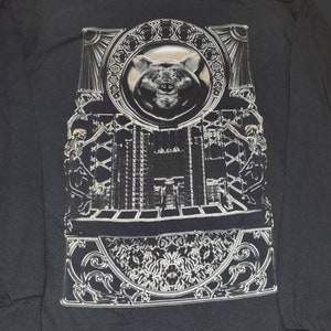 May include: Black long-sleeve shirt with a silver graphic print of a bear's head inside a circular frame with ornate details. The bear's head is surrounded by a skeletal figure on each side.