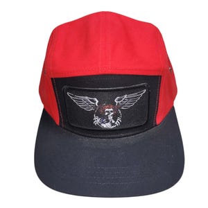 May include: A five-panel cap featuring a bright red crown and a solid black flat brim. The front panel displays a rectangular black patch with an embroidered graphic of a skull with wings in white and red thread, centered on the front of the hat.