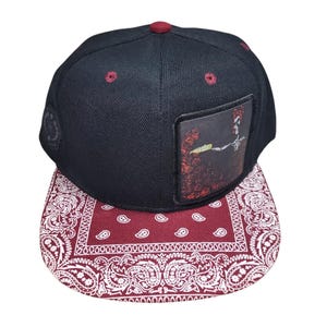 May include: A black baseball cap featuring a maroon and white paisley patterned flat brim. The front panel displays a square patch with a dark graphic illustration, while the crown is accented with maroon eyelets and a matching top button.