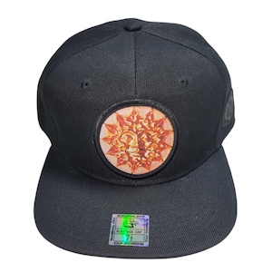 May include: Black flat-bill cap with a circular patch featuring a sun design in orange and gold. The cap has a structured fit and a flat brim. A holographic sticker is affixed to the brim.