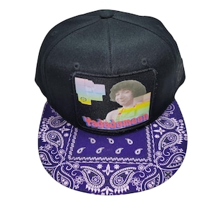 May include: A black baseball cap with a flat brim featuring a purple and white paisley pattern. The front panel displays a rectangular patch with a graphic of a person with an afro and the text "Yadadamean" in colorful, stylized lettering.