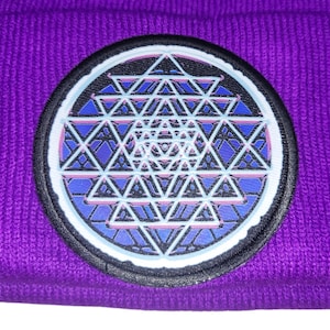 Op de afbeelding: Een paarse beanie met een cirkelvormige patch met een geometrisch ontwerp. De patch heeft een zwarte rand, een witte binnenring en een blauw en wit geometrisch patroon. De beanie is gemaakt van gebreid materiaal.