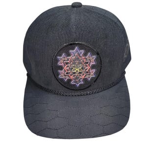 May include: Black corduroy baseball cap with a geometric design on the bill. The cap features a round patch with a colorful, intricate geometric pattern. The cap has a curved brim and a button on top.