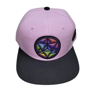 May include: A lavender and black baseball cap with a geometric design. The cap has a lavender crown, a black brim, and a circular patch with a colourful geometric pattern. The cap has a black button on top.