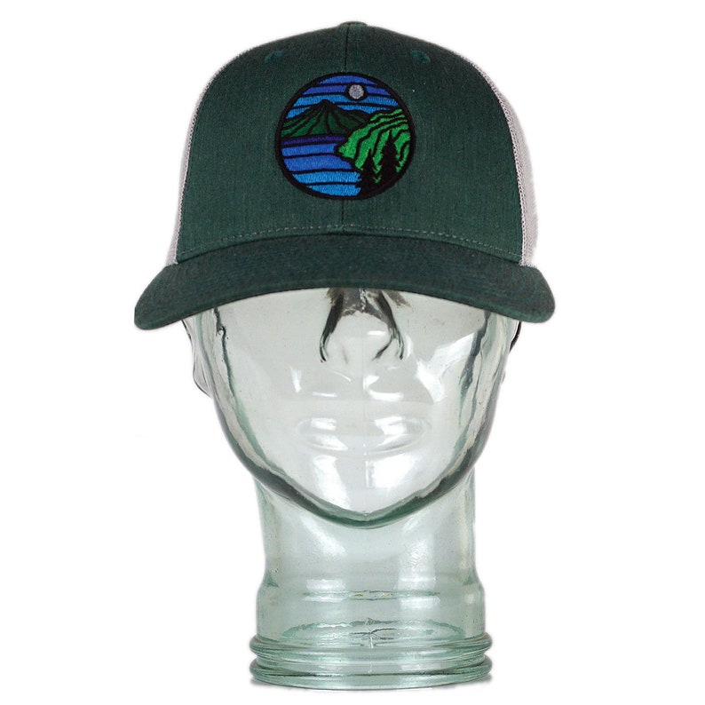 May include: A green and white trucker hat with a circular embroidered patch featuring a mountain scene with a lake and a moon.