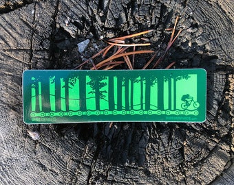 May include: A rectangular green sticker featuring a silhouette of a forest and a mountain biker riding along a bicycle chain. The text RISE DESIGNS is printed in the bottom left corner and RISERGRAPHICS.COM is printed in the bottom right corner.