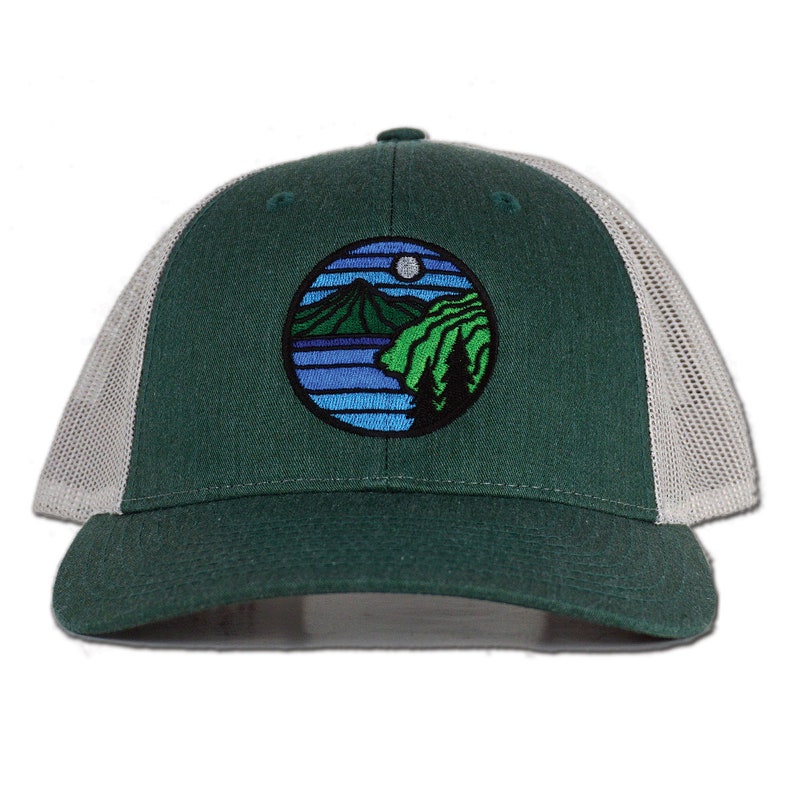 May include: A green and white trucker hat with a circular embroidered patch featuring a mountain scene with a moon and stars. The patch is blue, green, and white.