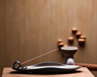 May include: A black ceramic incense holder with a curved design, holding a lit incense stick. The holder sits on a wooden table, with a blurred background featuring decorative elements and a warm color palette.