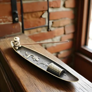 May include: A bronze-toned incense holder with a detailed design. The holder features a small figure, koi fish, and a cylindrical element. An incense stick is placed in the holder, resting on a wooden surface, with a brick wall in the background.