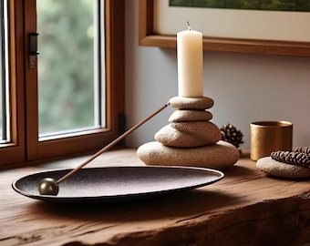 May include: A serene display with a white candle on a pile of stones. A lit incense stick rests in a dark oval dish with a gold sphere. A small gold cup and pine cones are also present, all arranged on a wooden surface, creating a calming atmosphere.