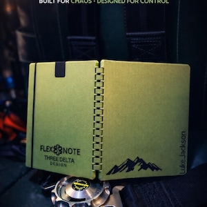 Può includere: Un taccuino Flexnote verde con dorso nero, con il testo "FLEXNOTE" e "BUILT FOR CHAOS • DESIGNED FOR CONTROL". Il taccuino presenta una grafica di montagna e il testo "THREE DELTA DESIGN."