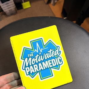Puede incluir: Un cuaderno rectangular de color amarillo neón brillante con un emblema azul de la Estrella de la Vida y una línea de latido blanca. El texto "THE MOTIVATED PARAMEDIC" está impreso en letras blancas y azules sobre el centro del emblema.