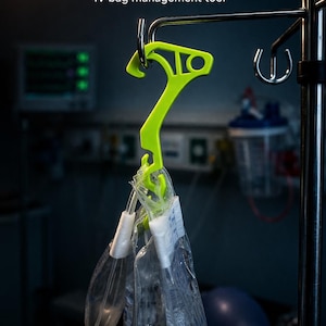 Puede incluir: Una herramienta de gestión de bolsas intravenosas de color verde lima, etiquetada como "DRIPHOOK", cuelga de un poste de metal. Bolsas intravenosas transparentes cuelgan del gancho. El fondo es un entorno médico borroso.