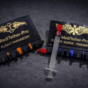 May include: Two black MediTether-Pro cases with gold lettering and a medical emblem. One case has orange accents, the other blue. A syringe and two red-capped tubes are also visible. The text on the cases reads "Flight Paramedic" and "Flight Nurse / Paramedic."