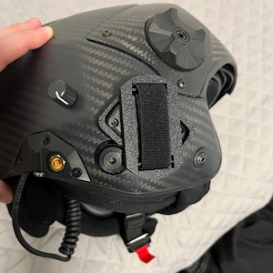 May include: A black carbon fiber helmet with a black strap and a red buckle. The helmet has a gold-colored antenna and a black knob on the side.