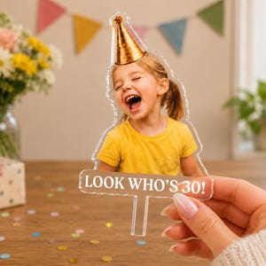 May include: A clear acrylic cake topper featuring a cutout of a laughing child wearing a gold party hat, with the white text LOOK WHO'S 30! printed on a rectangular base. The topper has a single stake at the bottom for insertion into a cake.