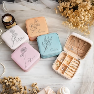 Może przedstawiać: Kolekcja spersonalizowanych pudełek na biżuterię w różnych pastelowych kolorach, w tym białym, brzoskwiniowym, niebieskim i różowym. Każde pudełko ma kwiatowy wzór i imię. Jedno pudełko jest otwarte, ukazując przegródki na biżuterię.