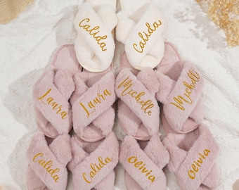 Può includere: Una collezione di pantofole in morbido tessuto con fasce incrociate, bianche e rosa chiaro. Ogni pantofola presenta un nome ricamato in oro: Calida, Laura, Michelle e Olivia. Le pantofole sono disposte su una superficie bianca.