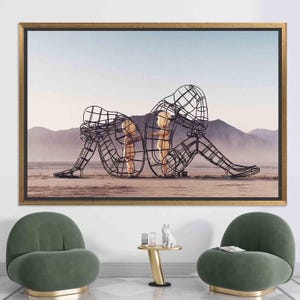 May include: A black and white photograph of three wire frame sculptures in a desert landscape. The sculptures are of two children and an adult, all standing in a line. The adult sculpture is sitting down with their head resting on their hand. The children are standing next to each other, facing the adult.