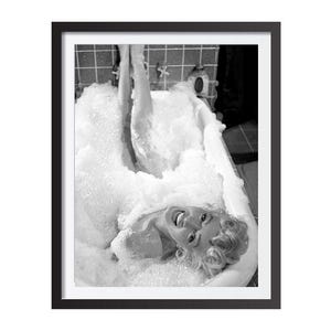 May include: A black and white photo of a person in a bathtub full of bubbles. The person is smiling and looking up at the camera. The photo is framed in a black frame.