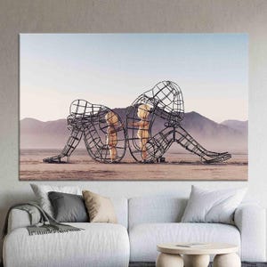 May include: A black metal sculpture of two figures, one larger than the other, standing in a desert landscape. The figures are made of wire mesh and appear to be embracing.