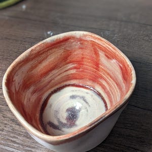 May include: A small, square ceramic bowl with a white glaze and red and brown swirling patterns inside.