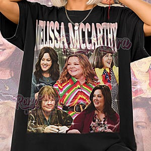 May include: Black unisex t-shirt featuring a graphic of Melissa McCarthy with the text "MELISSA MCCARTHY" above the image. The shirt also has the text "Unisex Shirt" at the bottom.