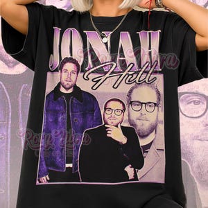 May include: Black unisex t-shirt featuring a vintage-style graphic of Jonah Hill. The design includes the name "Jonah Hill" in a purple, retro font, along with three images of the actor. The words "Unisex Shirt" are printed at the bottom.