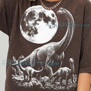 Peut inclure: T-shirt marron foncé avec un motif de dinosaures et de lune. Le design présente trois dinosaures et une pleine lune. Le texte "COMFORT COLORS ESPRESSO" est en bas. Le t-shirt est de couleur espresso, avec une impression de style vintage.