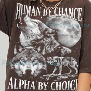 May include: Espresso-colored t-shirt featuring a wolf design with a full moon backdrop. The shirt has the phrases "HUMAN BY CHANCE" and "ALPHA BY CHOICE" printed on it. The shirt is made by Comfort Colors.