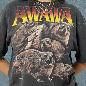 Könnte beinhalten: Dunkelgraues T-Shirt mit einem Aufdruck von Klippschliefern und dem Wort "AWAWA" in Gelb und Rot. Das T-Shirt trägt auch die Aufschrift "COMFORT COLORS" und "PEPPER" in Lila. Das Design ist auf einem Comfort Colors T-Shirt gedruckt.