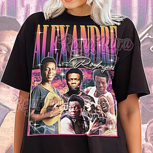 May include: Black unisex t-shirt featuring a colorful graphic design with the text "ALEXANDRE Rodrigues" and images of several people. The shirt also has the text "Unisex Shirt" at the bottom.