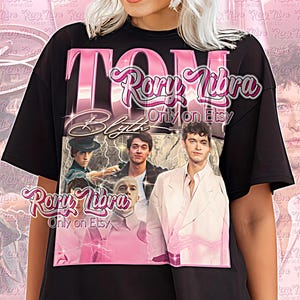 May include: Black unisex shirt with a pink graphic featuring the text "TOM Blyth" and the words "Rory Libra Only on Etsy." The graphic includes images of actors. The shirt also has the text "Unisex Shirt."