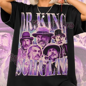 Pode incluir: Camiseta unissex preta com um gráfico roxo de cinco homens de chapéu e o texto "DR. KING SCHULM". A camisa também tem o texto "Unisex Shirt" em rosa.