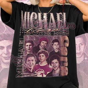 May include: Black unisex t-shirt featuring a graphic of a group of men with the text "MICHAEL CERA" in a vintage style. The shirt also has the words "Unisex Shirt" in pink script.