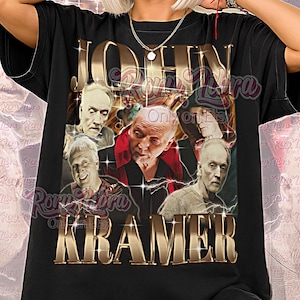 Könnte beinhalten: Schwarzes Unisex-T-Shirt mit einem goldfarbenen Design mit dem Namen "JOHN KRAMER" und Bildern von Männern. Die Worte "Unisex Shirt" sind unten auf dem Shirt in Rosa aufgedruckt.