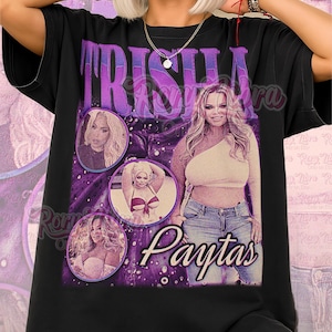 May include: Black unisex t-shirt featuring a graphic design with the name "Trisha" in purple, along with multiple images of a woman. The word "Paytas" is also visible. The shirt has the text "Unisex Shirt" in pink.