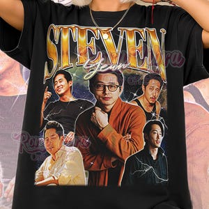 May include: Black unisex shirt featuring a graphic of Steven Yeun with the word "STEVEN" in gold lettering. The shirt has multiple images of Steven Yeun. The words "Unisex Shirt" are printed at the bottom in pink and white.