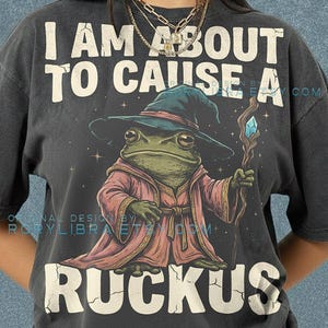 May include: Dark grey t-shirt with the text "I AM ABOUT TO CAUSE A RUCKUS." The shirt features a cartoon frog wizard with a staff. The frog is wearing a hat and robe. The shirt is Comfort Colors brand.