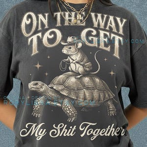 Peut inclure: T-shirt gris foncé avec un motif d'une souris portant un chapeau de cowboy sur une tortue, avec le texte "ON THE WAY TO GET My Shit Together". Le t-shirt est en tissu Comfort Colors Pepper.