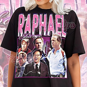 May include: Black unisex shirt featuring a graphic of the word "Raphael Sbarge" in pink, with a collage of images of people. The shirt also has the words "Unisex Shirt" in pink.