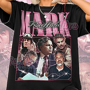 May include: Black unisex t-shirt featuring a pink graphic with the text "MARK Ruffalo" and images of the actor. The shirt has a vintage aesthetic and the words "Unisex Shirt" are printed at the bottom.